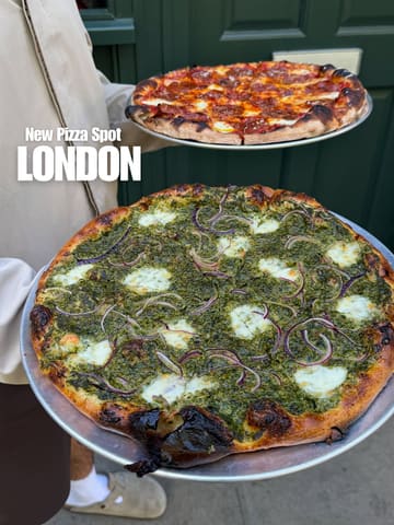 A casual, eye-level shot of two pizzas being held in front of a green door, likely outside a restaurant. The pizzas appear to be a pepperoni option and a green-topped vegetable option.