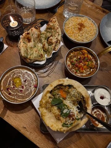 A top-down, overhead shot of a table spread at an Indian restaurant, featuring a variety of dishes including biryani, dal makhani, paneer lababdar, and naan, accompanied by a candle and glassware.