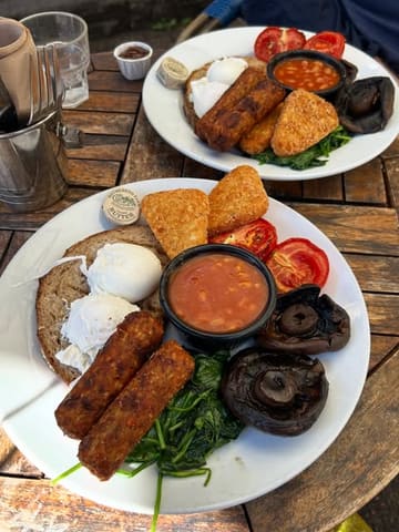 A top-down, flat-lay shot of two vegetarian breakfast plates served on a wooden outdoor table, featuring poached eggs, hash browns, veggie sausages, mushrooms, beans, spinach, and sourdough toast.