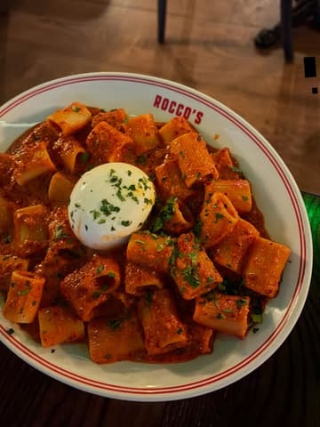 A top-down, appetizing shot of a plate of rigatoni pasta in a rich red sauce, topped with a whole ball of burrata cheese, served at Rocco's.