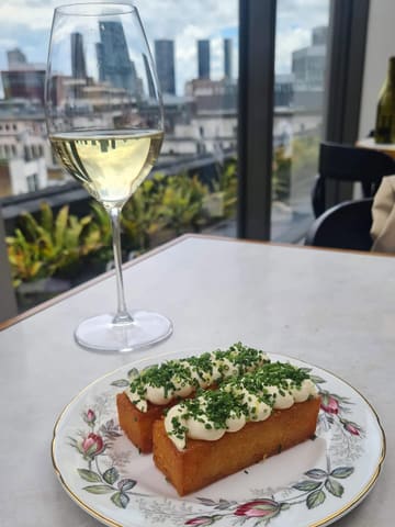 A high-quality, well-composed shot of a signature hash brown dish topped with taramasalata and chives, accompanied by a glass of white wine, set against a Manchester city skyline backdrop.