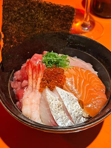 A close-up, overhead shot of a chirashi sushi bowl featuring various types of sashimi, shrimp, and roe, served in a dark bowl with a sheet of nori.
