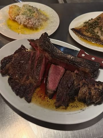 A high-angle, close-up shot of a medium-rare rib-eye steak served on a stainless steel counter, accompanied by side dishes, capturing a rustic, high-quality dining experience.