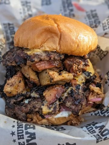 A high-quality, close-up shot of a 'Hungry Cowboy' burger, featuring smashed beef patties, smoked pork, and melted cheese, served on branded paper. The image captures the texture and 'dirty' aesthetic of the street food style perfectly.