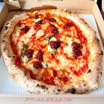 A high-quality, top-down photograph of a takeaway pizza from Rudy's, showcasing the melted cheese, tomato sauce, and toppings like nduja and artichokes inside the cardboard delivery box.
