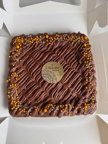 A high-angle, well-lit shot of a square brownie slab presented in a white takeaway box. The brownie is decorated with a neat chocolate drizzle and gold and silver sprinkles, featuring a central 'Ramadan Kareem' topper.