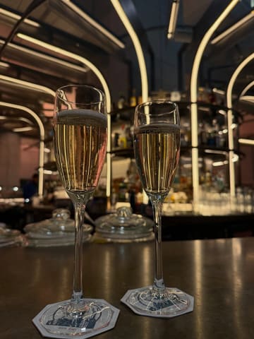 A high-quality, atmospheric shot of two glasses of champagne sitting on a bar counter, with the blurred, warm-lit interior of a bar in the background. The image captures a sophisticated, relaxing vibe, likely at a cocktail bar or lounge.