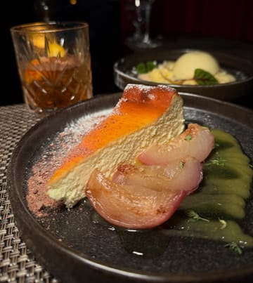 A high-quality, moody food photograph featuring a slice of basque cheesecake with poached fruit, served in a restaurant setting.