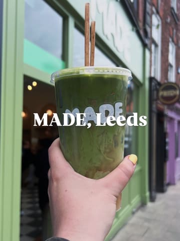 A first-person perspective shot holding a plastic takeaway cup containing a layered drink (half matcha, half iced coffee) in front of a green-fronted cafe in Leeds.