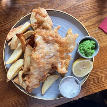 A top-down, casual shot of a classic fish and chips meal served on a wooden table. The image captures the texture of the battered fish and the accompanying sides, including minted peas and tartar sauce, typical of a British pub setting.