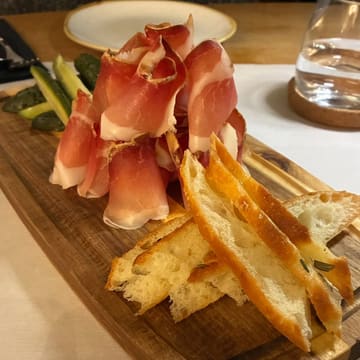 A close-up shot of a charcuterie sharing board featuring thinly sliced speck, pickled cucumbers, and toasted focaccia. The lighting is warm and appropriate for the setting, though the composition suffers from tight cropping.