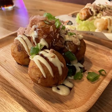 A close-up, high-quality shot of a serving of takoyaki on a wooden tray, topped with bonito flakes, mayonnaise, and green onions. The background is softly blurred, showing hints of other dishes and the restaurant environment.