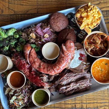 A top-down, flat-lay shot of a large BBQ sharing platter featuring a variety of smoked meats, sides, and sauces. The image effectively showcases the abundance and variety of the meal, typical of a casual BBQ restaurant setting.