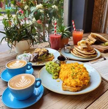 A bright, well-composed photograph of a brunch spread at a cafe, featuring scrambled eggs on toast, avocado toast, pancakes, and flat white coffees. The natural lighting and clean table setting create an inviting atmosphere.