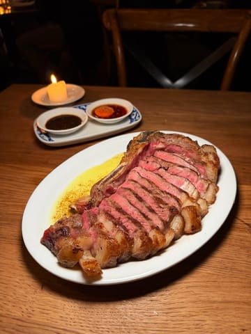 A high-quality, moody food photograph featuring a perfectly cooked, sliced bone-in sirloin steak served with dipping sauces in a dimly lit restaurant setting. The image effectively uses shallow depth of field to draw attention to the texture and doneness of the meat, creating an inviting and sophisticated atmosphere.