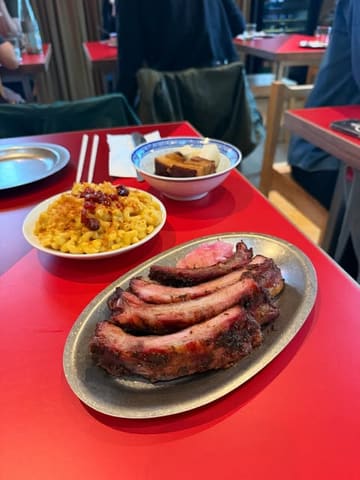 A top-down, slightly angled shot of a BBQ meal featuring pork ribs, mac and cheese, and pork belly, served on a bright red table in a restaurant setting.