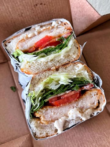 A close-up, high-quality shot of a chicken caesar sandwich served in a takeaway box. The image highlights the fresh ingredients, including the breaded chicken, crisp lettuce, tomato, and generous amount of dressing, all held within slices of focaccia.