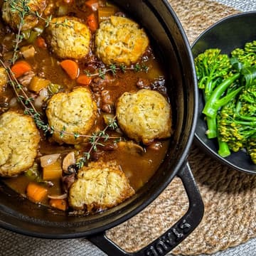 A high-quality, appetizing close-up shot of a homemade beef stew with dumplings in a cast-iron pot, served with a side of broccolini.