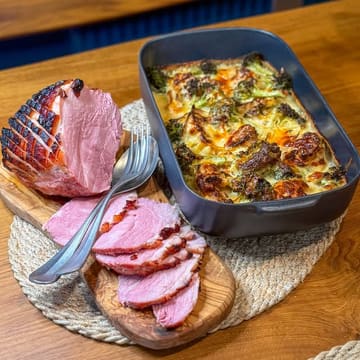 A high-quality, well-lit food photography shot featuring a glazed roast ham alongside a bubbling three-cheese cauliflower and broccoli bake, presented on a wooden board and placemat.