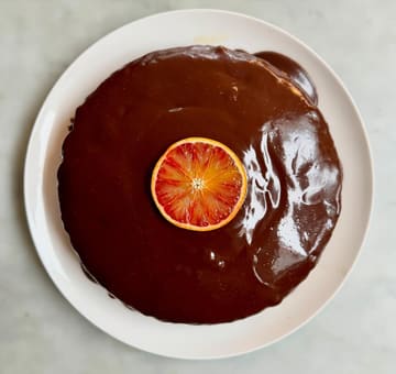 A high-angle, top-down shot of a round cake covered in a glossy chocolate ganache, topped with a single slice of blood orange. The cake is presented on a white plate against a neutral, light-colored marble surface, creating a clean and minimalist aesthetic.