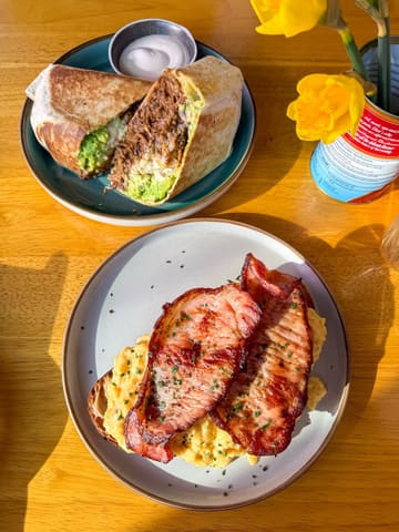 A bright, natural-light photograph of a breakfast spread featuring a burrito and scrambled eggs on sourdough, set on a wooden table with a small vase of daffodils.