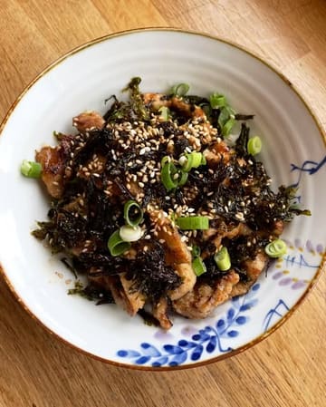 A simple, overhead shot of a pork rice bowl topped with seaweed, sesame seeds, and spring onions, served in a patterned ceramic bowl on a wooden table. The image captures a casual, home-style meal at a local cafe.