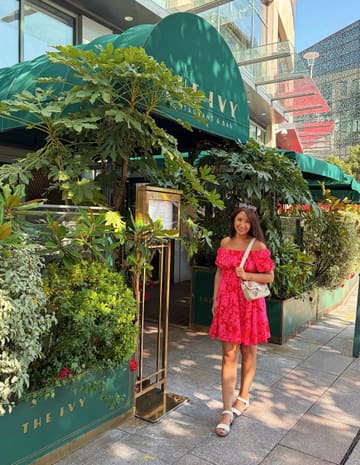 A bright, well-composed portrait of a woman standing in front of The Ivy restaurant in Cardiff, showcasing the venue's exterior branding and outdoor seating area.