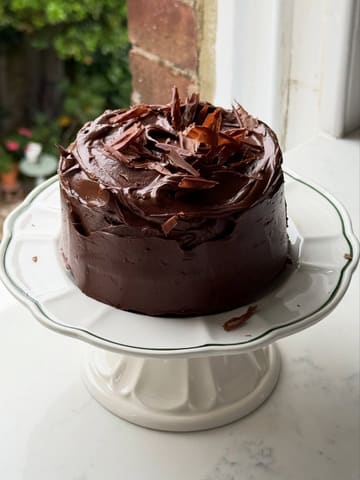 A high-quality, close-up photograph of a small, homemade chocolate fudge cake displayed on a white cake stand. The image utilizes soft, natural side-lighting to emphasize the texture of the chocolate buttercream frosting and the delicate chocolate curls on top, creating an inviting and professional aesthetic.