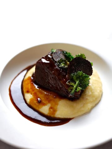 A high-quality, professional-looking food photograph of a braised short rib dish served on a bed of polenta, garnished with crispy kale and fresh truffle shavings. The image uses a clean, high-key aesthetic with a white background, emphasizing the rich, glossy texture of the meat and the elegant plating.