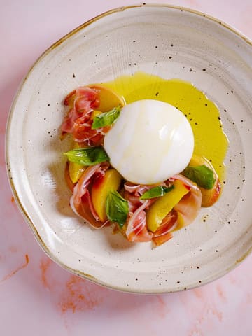 A high-quality, professional overhead food photograph featuring a burrata salad with roasted peaches, prosciutto di parma, and fresh basil, drizzled with olive oil on a textured ceramic plate.