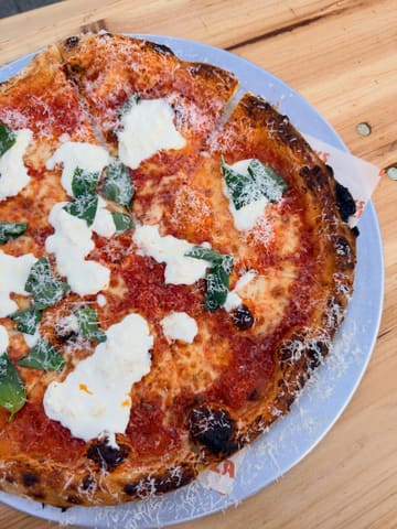 A top-down, natural-light shot of a Margherita pizza topped with stracciatella, served on a metal tray on a wooden table. The image captures the texture of the crust and the fresh ingredients, though the framing is quite tight.