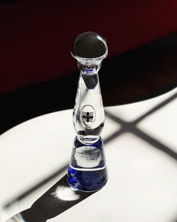 A high-quality, professional product shot of a Clase Azul tequila bottle placed on a marble surface. The lighting is dramatic and intentional, highlighting the unique shape and texture of the hand-blown ceramic decanter against a dark, moody background.