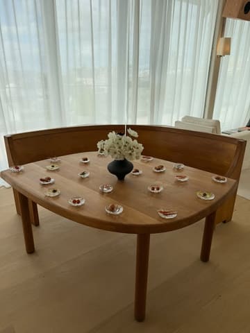 A minimalist, high-end restaurant interior shot featuring a wooden table set with sparse canapés and a central floral arrangement, bathed in bright natural light from a large window.