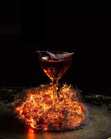 A highly stylized, editorial-quality photograph of a cocktail served in a coupe glass, surrounded by a dramatic, glowing pyrotechnic effect against a pitch-black background. The image emphasizes luxury and atmosphere, suitable for high-end hospitality marketing.