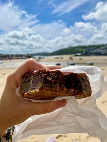 A close-up, handheld shot of a brookie (brownie-cookie hybrid) being held against a blurred beach and sea background.