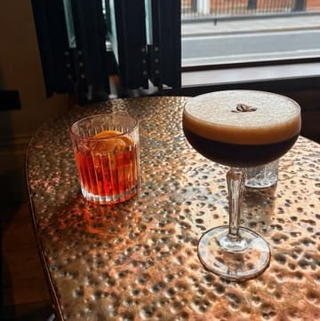 A high-quality, well-composed shot of two cocktails—an espresso martini and a negroni—resting on a textured copper table in a bar setting. The lighting is moody and atmospheric, highlighting the drinks effectively while maintaining a relaxed, intimate vibe suitable for a bar review.