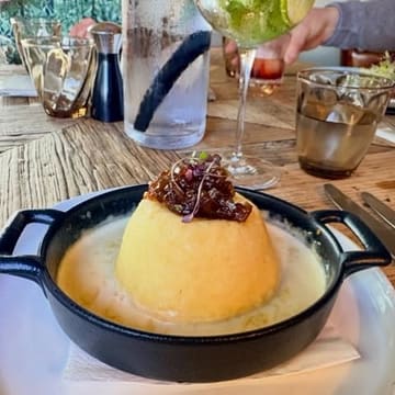 A close-up, slightly elevated shot of a twice-baked soufflé served in a black cast-iron dish, placed on a rustic wooden table. The background is cluttered with glassware, a water bottle, and a partial view of a person's arm.
