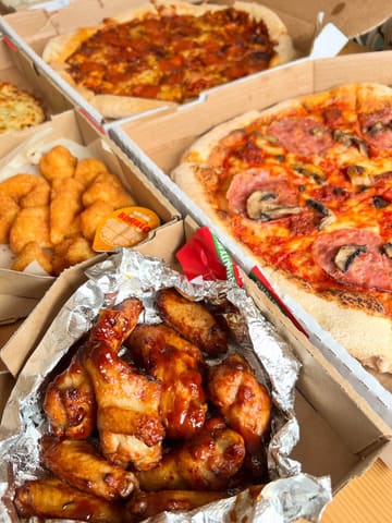 A flat-lay photograph of multiple Papa Johns takeaway boxes containing pizzas, chicken wings, and potato bites. The image captures a casual, at-home dining experience.