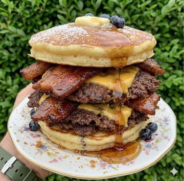 A close-up, vertical shot of a 'pancake burger' stack featuring pancakes, steak patties, bacon, and syrup, held by a hand against a hedge background.