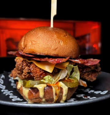 A close-up, high-contrast shot of a fried chicken burger featuring bacon, cheese, and pickles, set against a dark background. The lighting is punchy and direct, highlighting the textures of the fried chicken and the sauce drip.