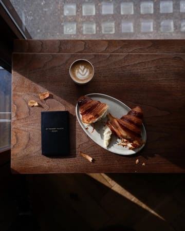 A high-quality, overhead flat-lay photograph featuring a flaky croissant, a latte with latte art, and a branded notebook on a wooden table, capturing a cozy, aesthetic bakery experience.