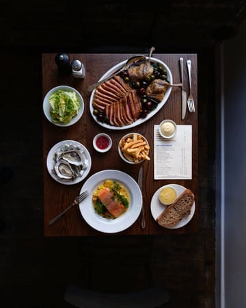 A high-quality, overhead flat-lay shot of a table spread at a restaurant, featuring a variety of dishes including duck, salmon, oysters, and fries, alongside a menu. The lighting is moody and atmospheric, highlighting the textures of the food.