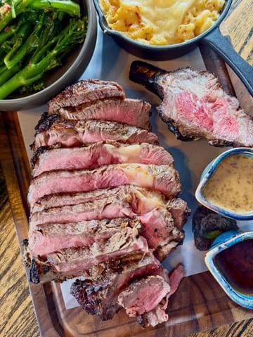 A high-angle, overhead shot of a premium gastropub meal featuring a sliced côte de boeuf steak, a cast-iron skillet of mac and cheese, broccolini, and two dipping sauces served on a wooden board.