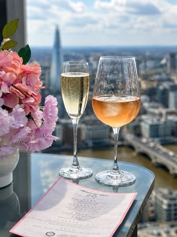 A high-quality, bright shot of two glasses of wine on a glass table, set against a stunning panoramic view of the London skyline, including The Shard. The scene is styled with a bouquet of pink flowers and a menu, creating an inviting, upscale atmosphere.