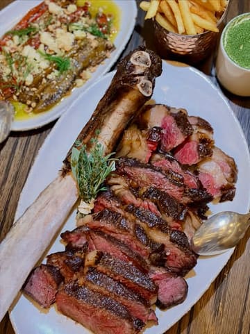 A high-quality, appetizing close-up shot of a 1.2kg Scottish Tomahawk steak served at a restaurant, accompanied by sides including fries and a prawn dish. The focus is sharp on the meat, highlighting the quality of the cook.