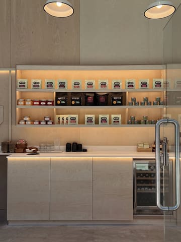 A clean, aesthetically pleasing, and well-lit photograph of a cafe's retail display shelves. The image showcases a variety of neatly arranged jars and bags of coffee or tea products against a minimalist plywood background, creating an inviting and professional atmosphere.