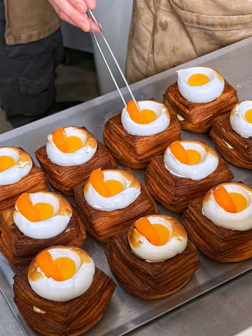 A high-quality, behind-the-scenes shot of a pastry chef finishing a batch of cube-shaped pastries topped with meringue and mandarin segments. The image captures the precision of the plating process in a clean, well-lit kitchen environment.