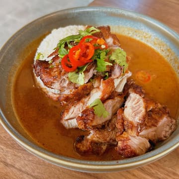 A high-quality, overhead shot of a Thai fried chicken dish served with rice and a generous portion of satay sauce, garnished with fresh coriander and sliced red chili.