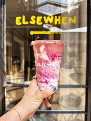 A vibrant, well-composed product shot of a smoothie held in front of the 'Elsewhen' shopfront, effectively using the brand's signage as a backdrop to establish the venue.