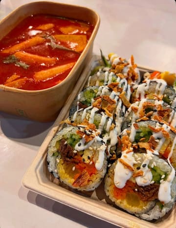 A close-up, high-angle shot of Korean street food, specifically beef kimbap and tteokbokki, served in takeaway containers. The lighting is bright and even, highlighting the textures of the food, though the composition feels slightly cramped within the frame.